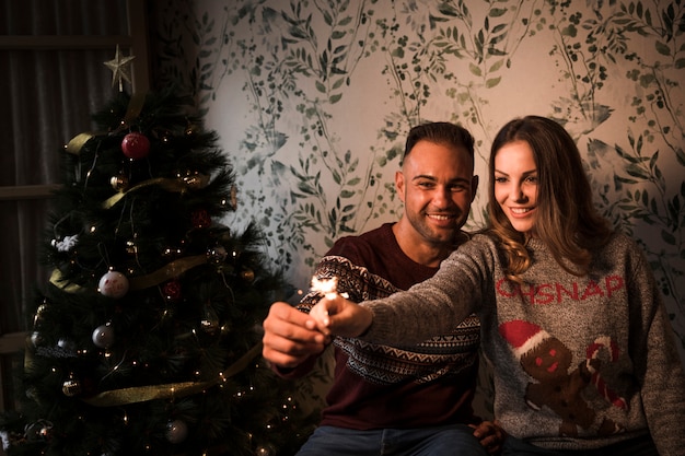 Foto gratuita chico joven cerca de una dama atractiva con luces de bengala cerca del árbol de navidad