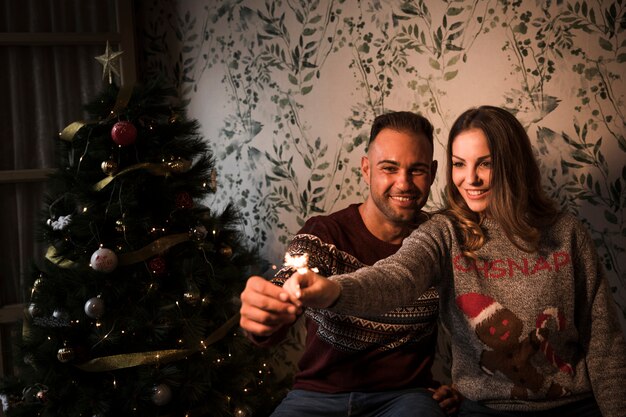 Chico joven cerca de una dama atractiva con luces de Bengala cerca del árbol de Navidad