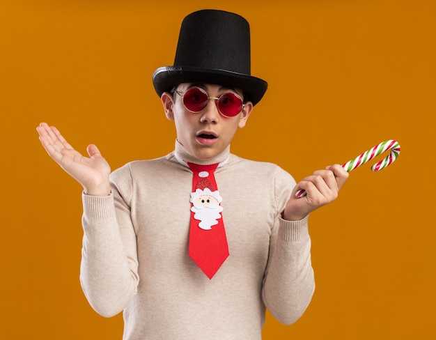 Foto gratuita chico joven confundido con sombrero con corbata navideña y gafas sosteniendo dulces navideños extendiendo la mano aislada en la pared amarilla