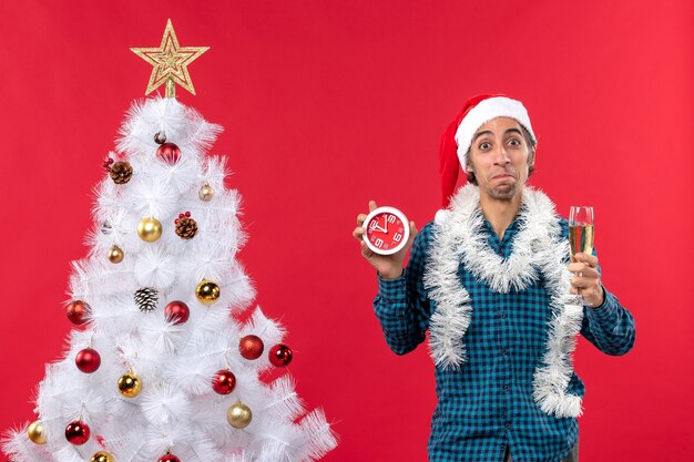 Chico joven curioso feliz con sombrero de santa claus y levantando una copa de vino y sosteniendo el reloj