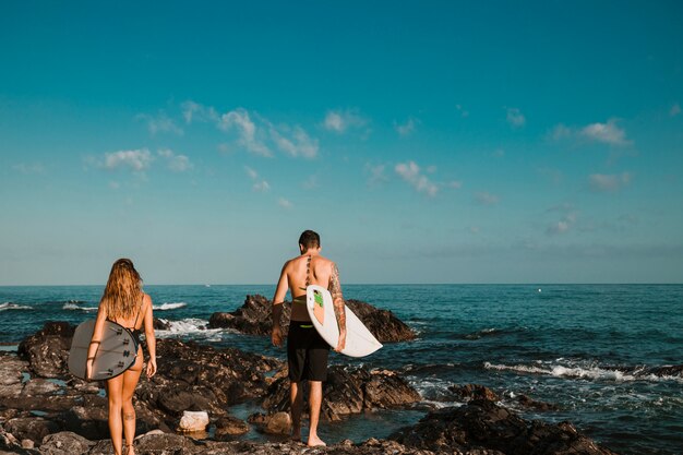 Chico joven y una dama con tablas de surf que van a la orilla de la piedra al agua