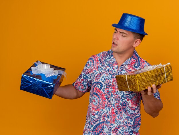 Chico joven disgustado con sombrero azul sosteniendo y mirando cajas de regalo aisladas en naranja