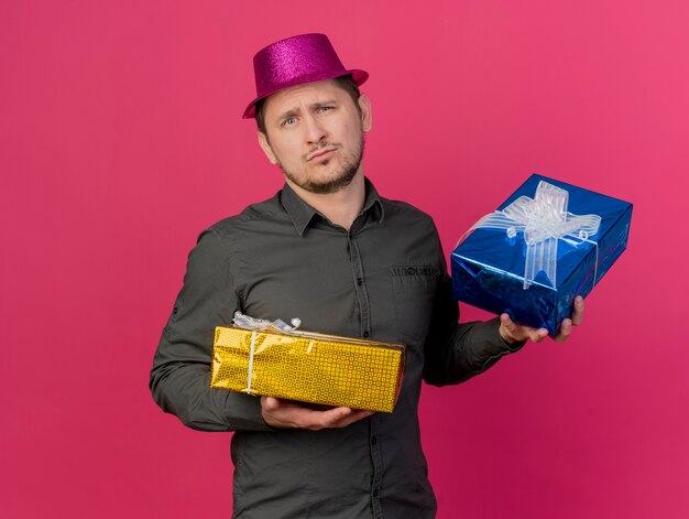 Chico joven disgustado con sombrero rosa sosteniendo cajas de regalo aisladas en rosa