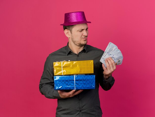 Chico joven disgustado con sombrero rosa sosteniendo cajas de regalo y mirando efectivo en su mano aislado en rosa