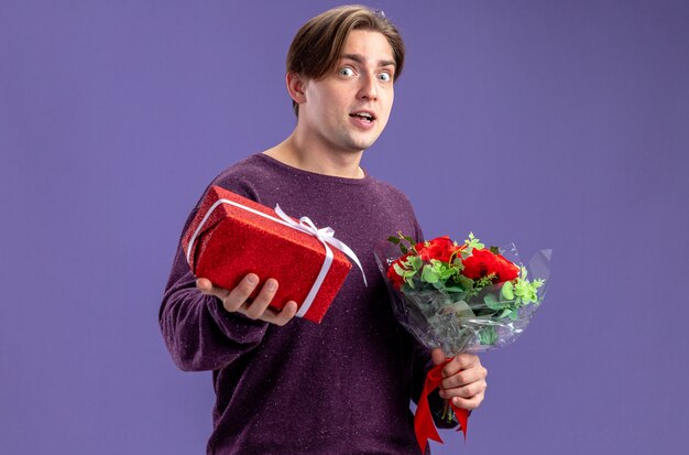 Chico joven emocionado en el día de San Valentín con caja de regalo con ramo aislado sobre fondo azul.