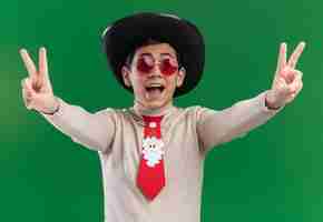 Foto gratuita chico joven emocionado con sombrero con corbata de navidad y gafas mostrando gesto de paz aislado sobre fondo verde