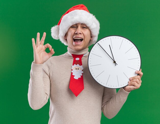 Chico joven emocionado con sombrero de navidad con corbata sosteniendo reloj de pared mostrando gesto bien aislado en la pared verde