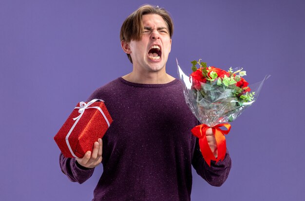 Chico joven enojado en el día de San Valentín con caja de regalo con ramo aislado sobre fondo azul.