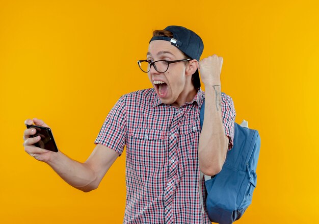 Chico joven estudiante alegre con mochila y gafas y gorra sosteniendo y mirando el teléfono mostrando gesto de sí