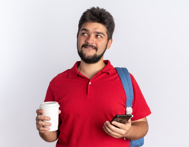 Chico joven estudiante barbudo en camisa polo roja con mochila sosteniendo teléfono inteligente y taza de café mirando a un lado con sonrisa astuta en la cara de pie sobre la pared blanca