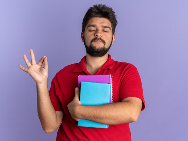 Chico joven estudiante barbudo en camisa polo roja sosteniendo libros haciendo gesto de meditación relajante con los ojos cerrados de pie sobre la pared azul