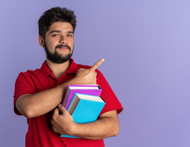 Chico joven estudiante barbudo en camisa polo roja sosteniendo libros mirando confiado sonriendo apuntando con el dedo índice al lado de pie