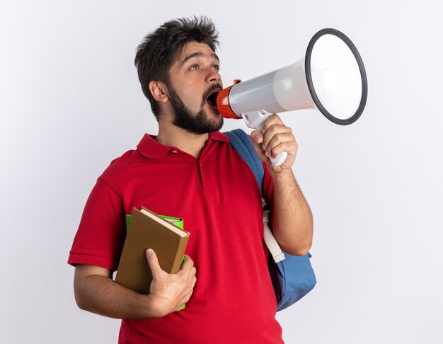 Chico joven estudiante barbudo emocional en polo rojo con mochila sosteniendo cuadernos gritando al megáfono de pie sobre la pared blanca