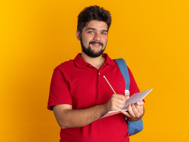 Chico joven estudiante barbudo feliz en polo rojo con mochila con cuaderno y lápiz escribiendo sonriendo alegremente de pie sobre la pared naranja