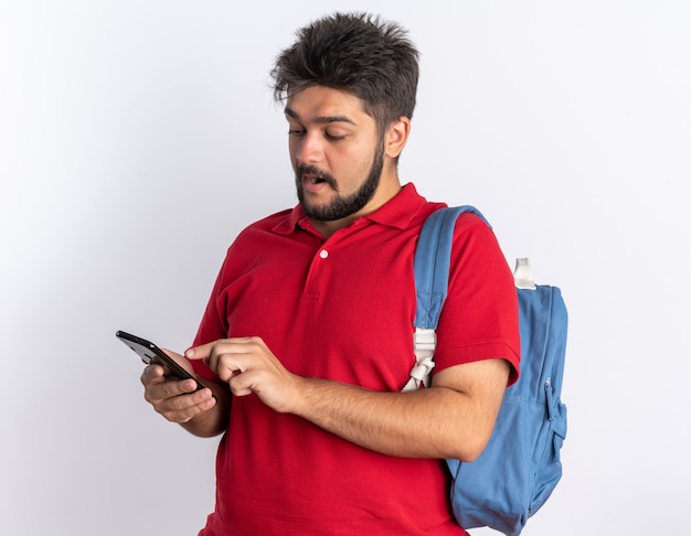 Chico joven estudiante barbudo feliz en polo rojo con mochila con smartphone escribiendo mensajes sonriendo de pie sobre fondo blanco.