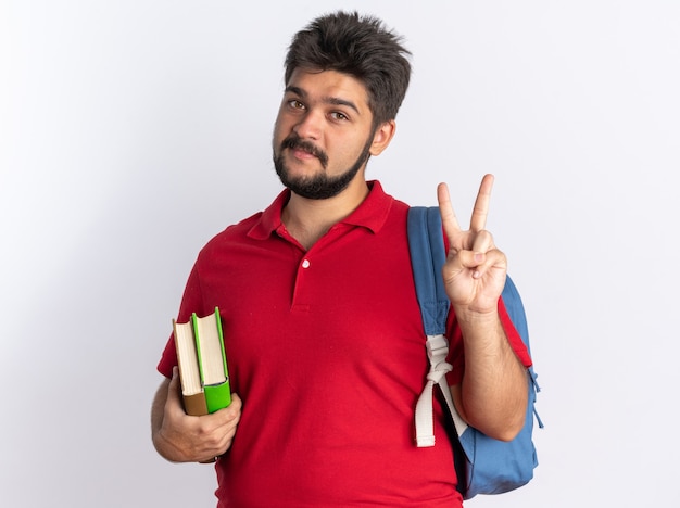 Chico joven estudiante barbudo feliz en polo rojo con mochila sosteniendo cuadernos mostrando signo v de pie sobre la pared blanca