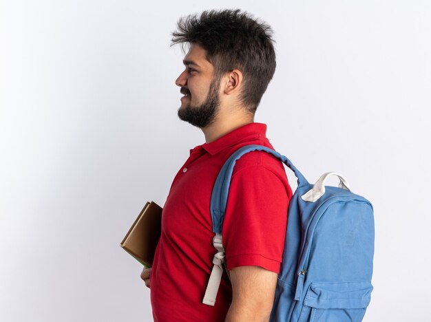 Chico joven estudiante barbudo feliz en polo rojo con mochila sosteniendo cuadernos de pie de lado sobre la pared blanca