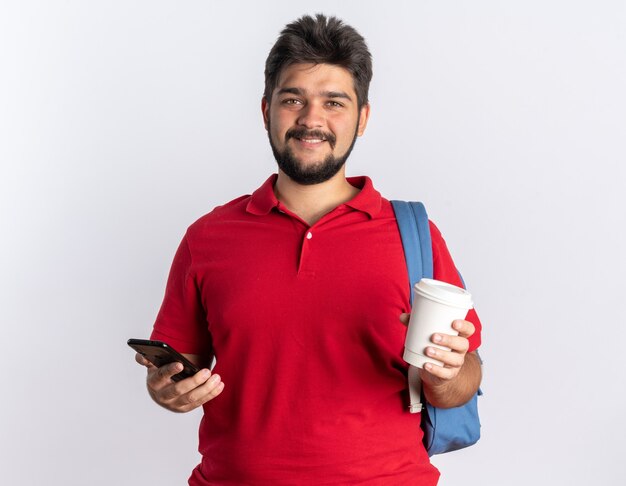 Chico joven estudiante barbudo feliz en polo rojo con mochila sosteniendo la taza de café y el teléfono inteligente sonriendo alegremente de pie sobre la pared blanca