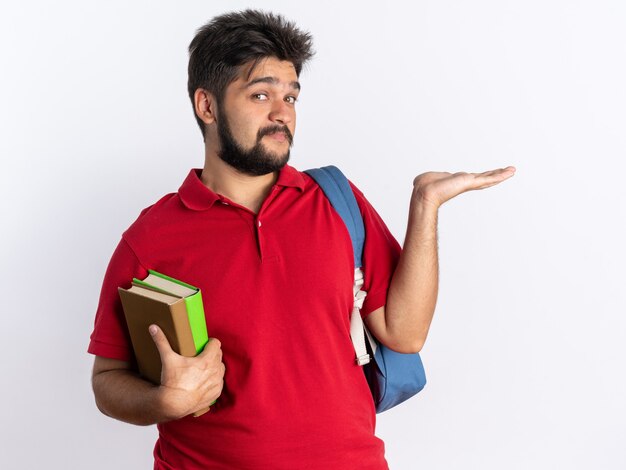 Chico joven estudiante barbudo feliz y positivo en polo rojo con mochila sosteniendo cuadernos que presentan espacio de copia con el brazo de su mano parado sobre la pared blanca