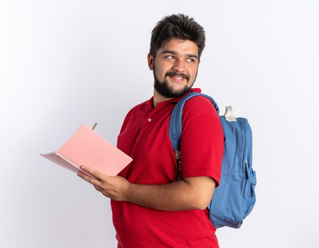Chico joven estudiante barbudo en polo rojo con mochila escribiendo en el cuaderno mirando confiado sonriendo de pie