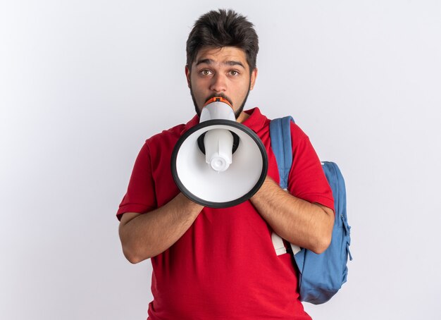 Chico joven estudiante barbudo en polo rojo con mochila gritando al megáfono siendo emocional y preocupado de pie