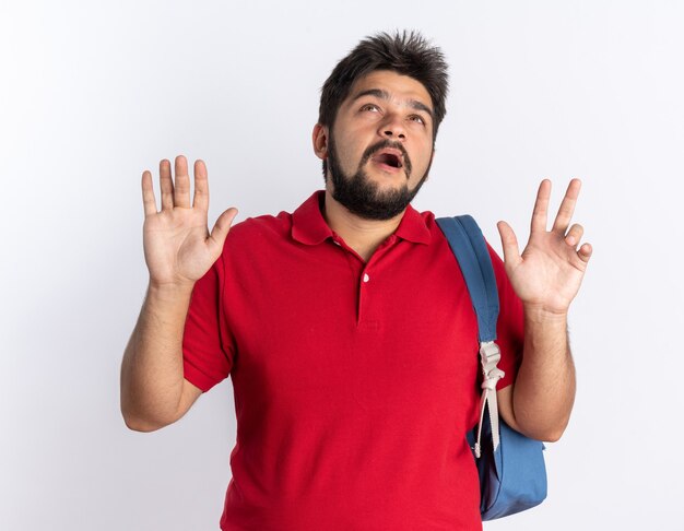 Chico joven estudiante barbudo en polo rojo con mochila mirando hacia arriba preocupado de pie sobre la pared blanca