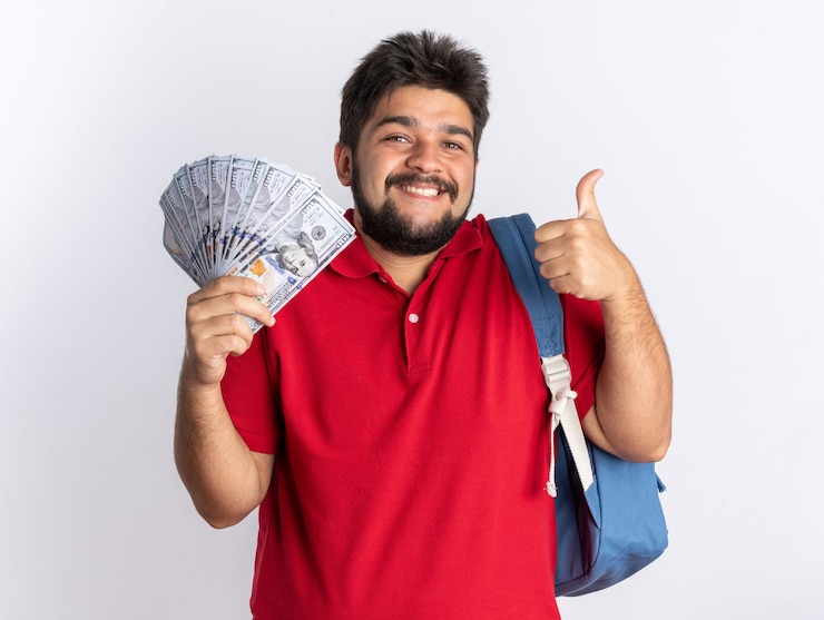 Foto gratuito chico joven estudiante barbudo en polo rojo con mochila sosteniendo boletos de avión y globo feliz y positivo mostrando los pulgares para arriba sonriendo de pie sobre la pared blanca