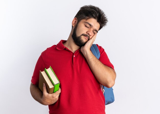 Chico joven estudiante barbudo en polo rojo con mochila sosteniendo cuadernos con aspecto cansado y aburrido, inclinando la cabeza en la palma de la mano sobre la pared blanca