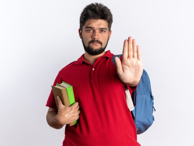 Chico joven estudiante barbudo en polo rojo con mochila sosteniendo cuadernos con cara seria haciendo gesto de parada con la mano de pie sobre la pared blanca