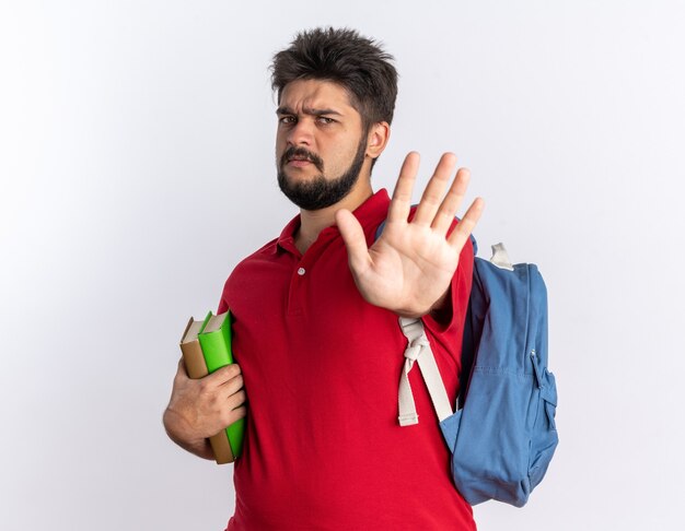 Chico joven estudiante barbudo en polo rojo con mochila sosteniendo cuadernos con el ceño fruncido haciendo gesto de parada con la mano de pie sobre la pared blanca