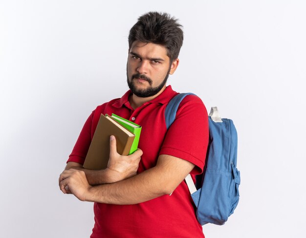 Chico joven estudiante barbudo en polo rojo con mochila sosteniendo cuadernos con el ceño fruncido de pie sobre la pared blanca