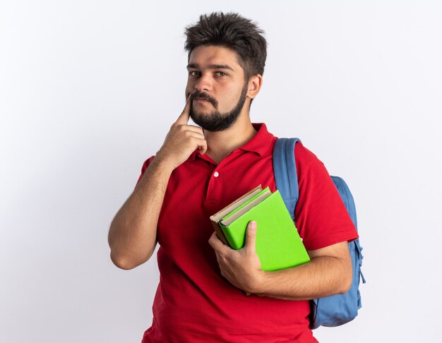 Chico joven estudiante barbudo en polo rojo con mochila sosteniendo cuadernos desconcertado de pie sobre la pared blanca