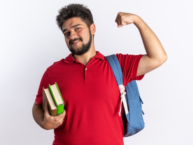 Chico joven estudiante barbudo en polo rojo con mochila sosteniendo cuadernos feliz y positivo mirando levantando puño sonriendo de pie