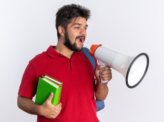 Chico joven estudiante barbudo en polo rojo con mochila sosteniendo cuadernos gritando al megáfono de pie feliz y positivo