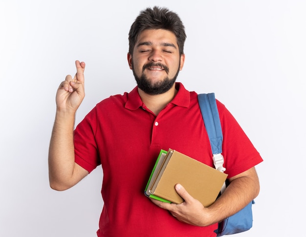 Chico joven estudiante barbudo en polo rojo con mochila sosteniendo cuadernos haciendo deseo cruzando los dedos con expresión de esperanza de pie