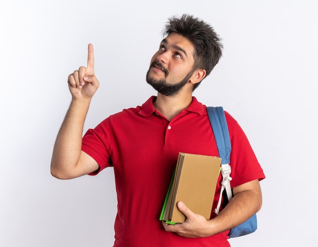 Chico joven estudiante barbudo en polo rojo con mochila sosteniendo cuadernos mirando hacia arriba con una sonrisa en la cara inteligente apuntando con el dedo índice a algo de pie