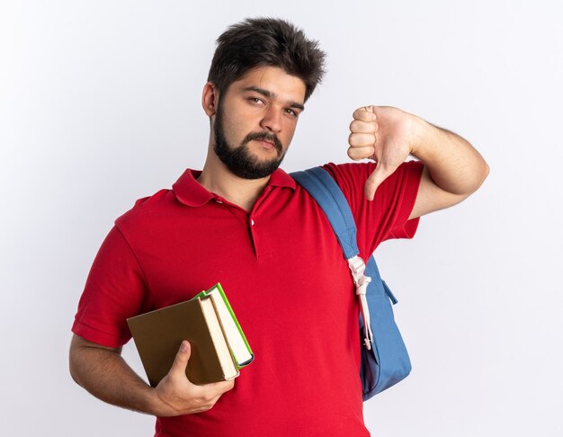 Chico joven estudiante barbudo en polo rojo con mochila sosteniendo cuadernos mirando estar disgustado mostrando los pulgares hacia abajo de pie