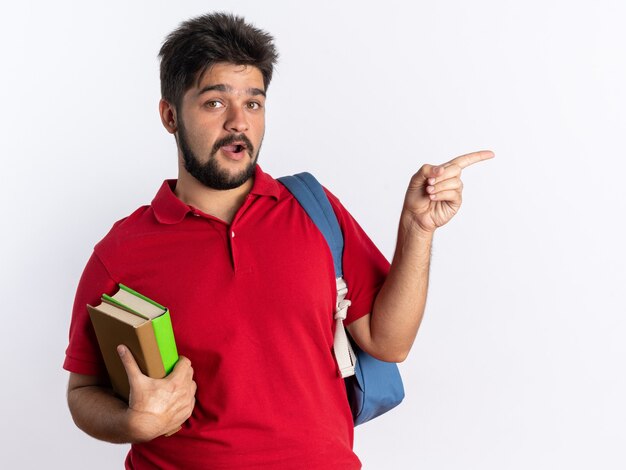 Chico joven estudiante barbudo en polo rojo con mochila sosteniendo cuadernos mirando feliz y sorprendido apuntando con el dedo índice al lado de pie