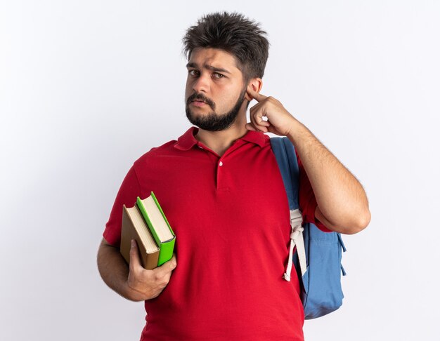 Chico joven estudiante barbudo en polo rojo con mochila sosteniendo cuadernos mirando intrigado con cara seria de pie