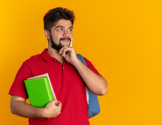 Chico joven estudiante barbudo en polo rojo con mochila sosteniendo cuadernos mirando a un lado con expresión pensativa pensando positivo sonriente de pie sobre fondo naranja