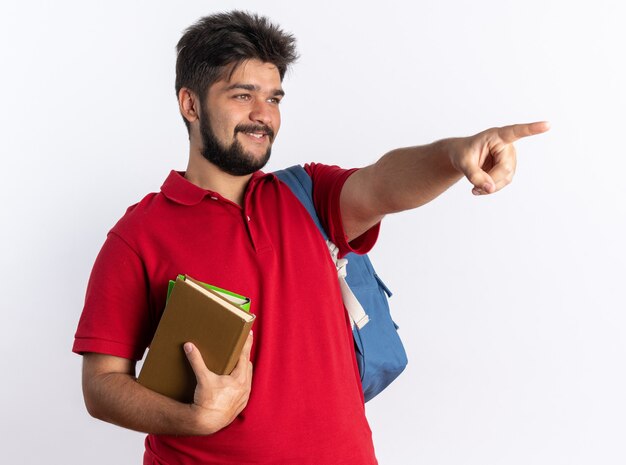 Chico joven estudiante barbudo en polo rojo con mochila sosteniendo cuadernos mirando a un lado sonriendo alegremente apuntando con el dedo índice a algo parado sobre la pared blanca