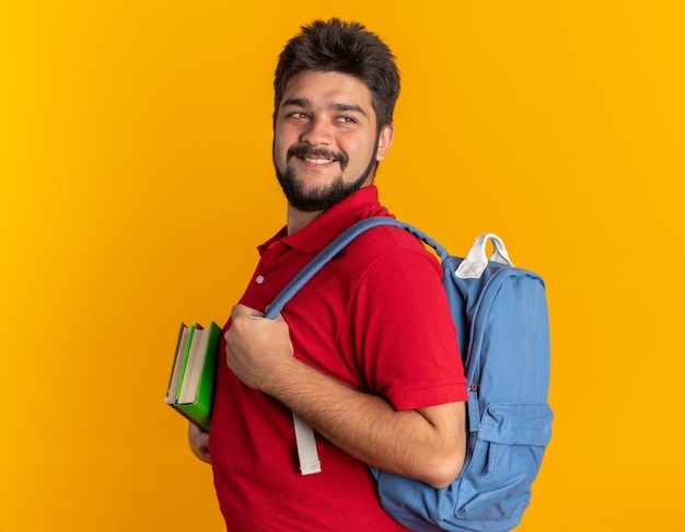 Chico joven estudiante barbudo en polo rojo con mochila sosteniendo cuadernos mirando a un lado sonriendo confiado, feliz y positivo de pie sobre la pared naranja