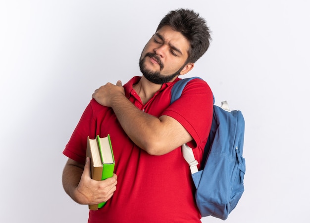 Chico joven estudiante barbudo en polo rojo con mochila sosteniendo cuadernos mirando mal tocando su hombro sintiendo dolor de pie