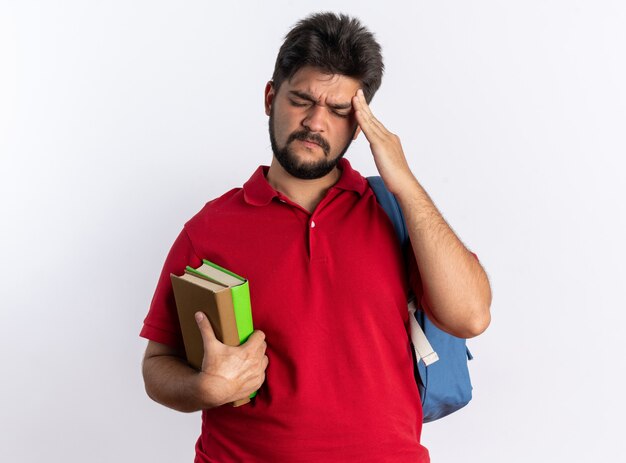 Chico joven estudiante barbudo en polo rojo con mochila sosteniendo cuadernos mirando mal tocando su sien sintiendo dolor de pie sobre una pared blanca