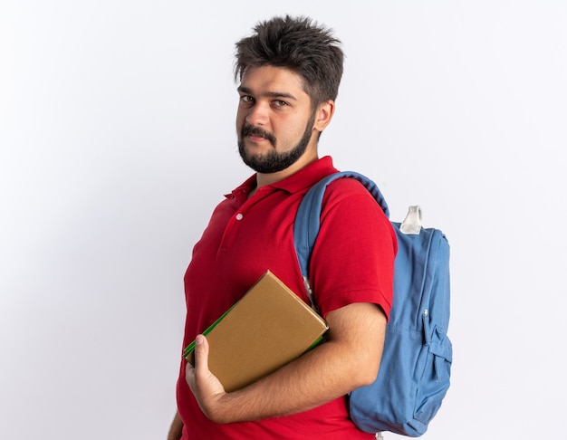 Chico joven estudiante barbudo en polo rojo con mochila sosteniendo cuadernos mirando sonriendo feliz y positivo de pie