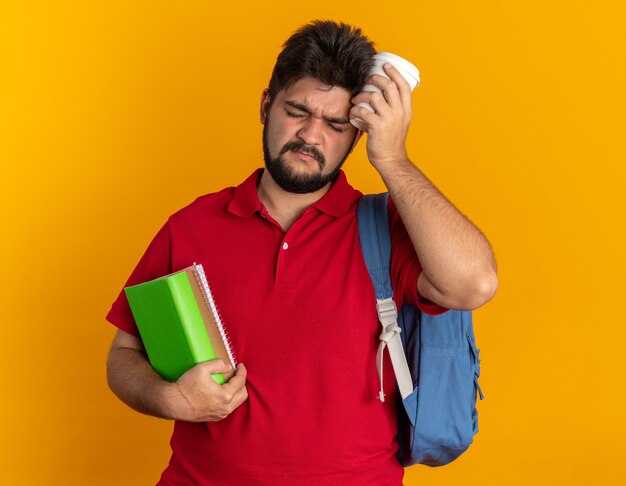 Chico joven estudiante barbudo en polo rojo con mochila sosteniendo cuadernos y taza de papel con aspecto cansado y con exceso de trabajo de pie sobre fondo naranja