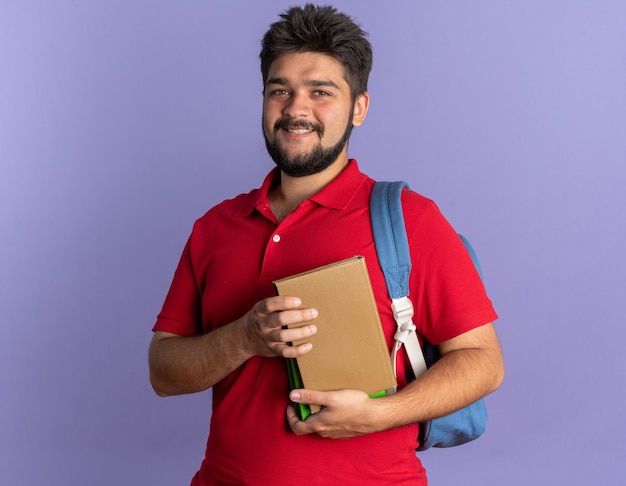 Chico joven estudiante barbudo en polo rojo con mochila sosteniendo libros mirando sonriendo alegremente feliz y positivo de pie