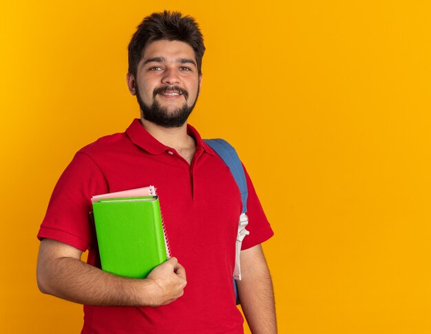 Chico joven estudiante barbudo en polo rojo con mochila sosteniendo libros sonriendo alegremente feliz y seguro de pie sobre la pared naranja