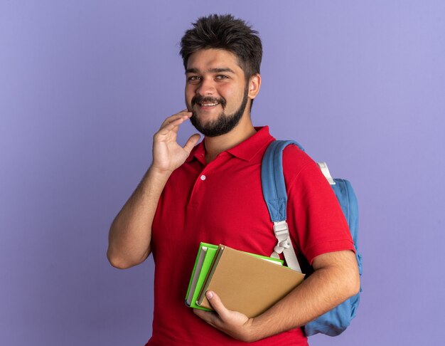 Chico joven estudiante barbudo en polo rojo con mochila sosteniendo libros con sonrisa tímida en la cara de pie sobre la pared azul