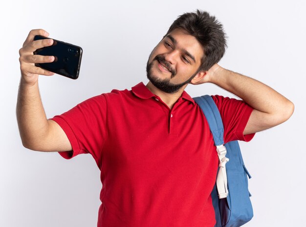 Chico joven estudiante barbudo en polo rojo con mochila sosteniendo smartphone haciendo selfie feliz y alegre sonriente de pie